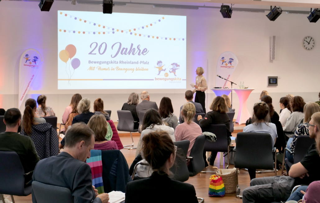Im Hintergrund eine Leinwand, auf der 20 Jahre Bewegungskita Rheinland-Pfalz steht, im Vordergrund sitzen viele Menschen