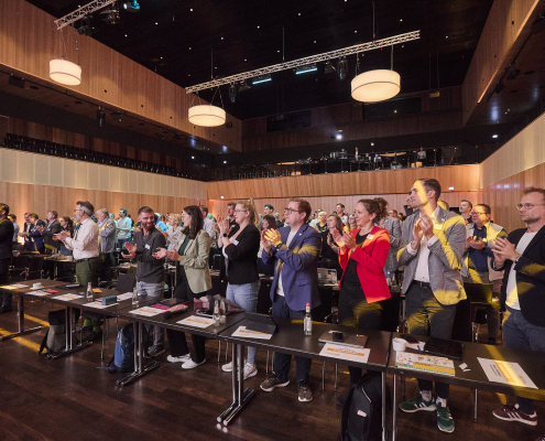 Personen stehen in Reihen in einem Saal hinter Tischen und applaudieren