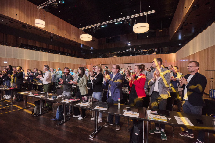 dsj-Hauptausschuss Personen stehen in Reihen in einem Saal hinter Tischen und applaudieren